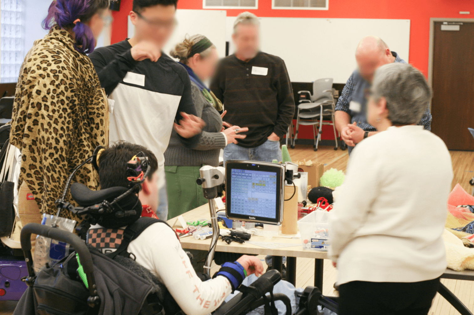 A photograph taken at a workshop with a person using an AAC device, her close conversational partner, and puppeteers. All participants stand around a table full of craft supplies and are engaged in coversation.